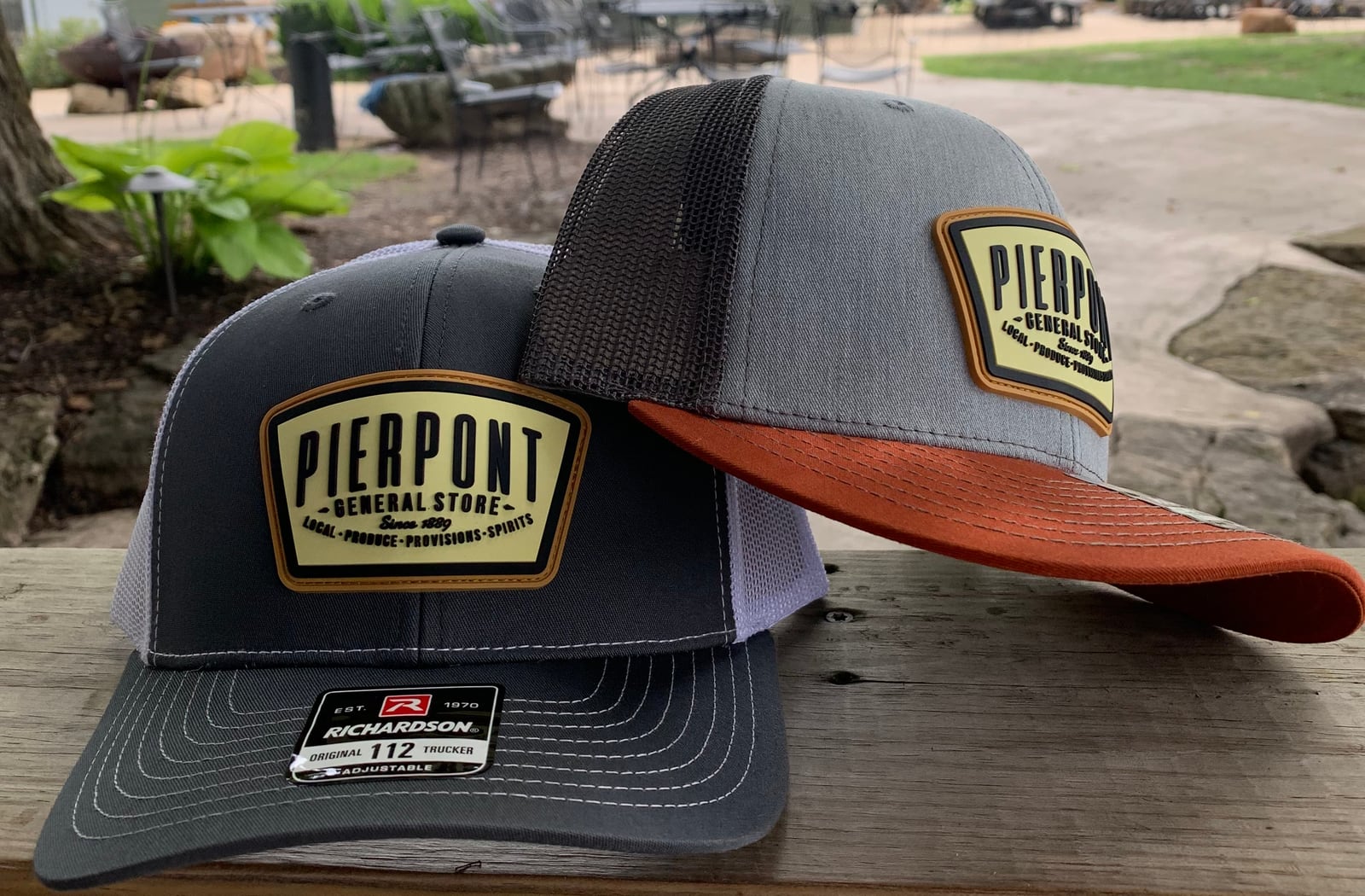 Two Pierpont General Store trucker hats in charcoal, gray and rust displayed outdoors