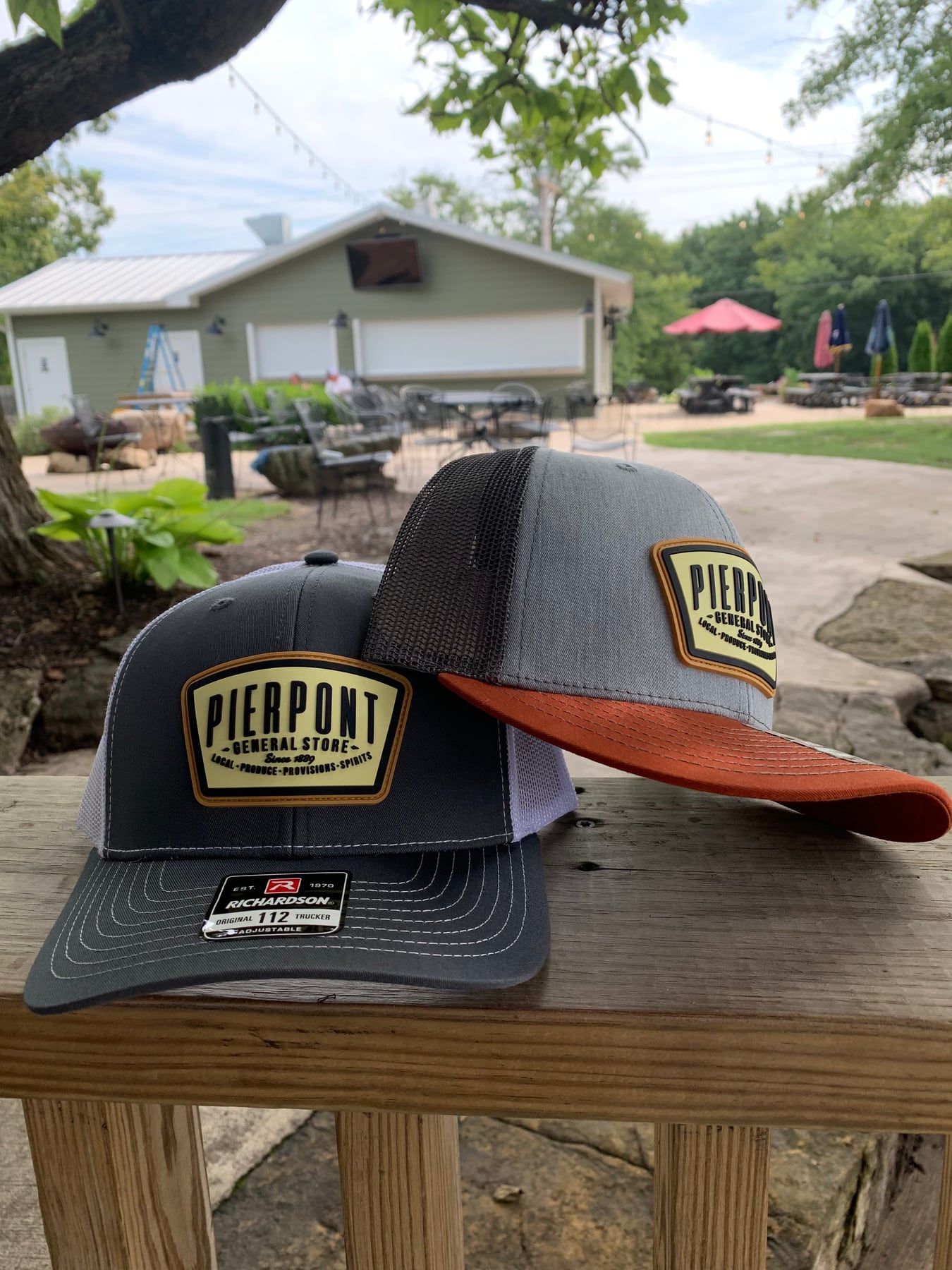 Two Pierpont General Store trucker hats in charcoal, gray and rust displayed outdoors